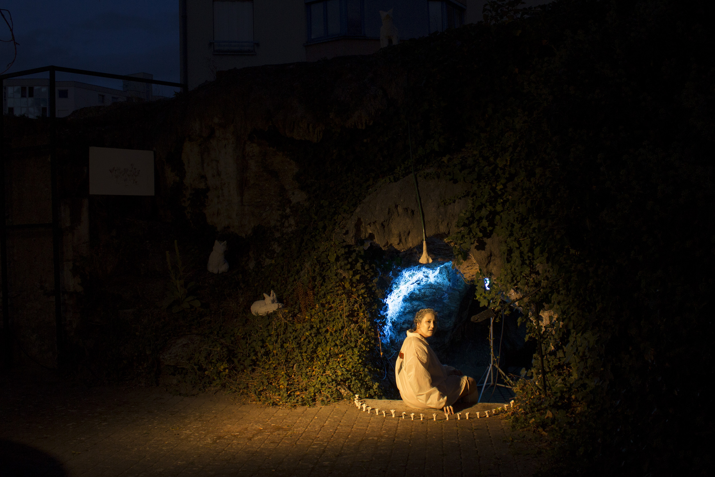 La photo est prise de nuit, on voit Clélia à l'entrée de la grotte qui est éclérée en bleu depuis l'intérieur. Un spot jaune délimite l'entrée de la grotte et éclaire Clélia. On voit le clercle de chanterelles et un renardeau.
